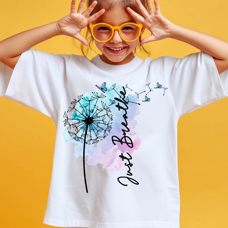 May include: White t-shirt featuring a dandelion design with blue butterflies and the words "Just Breathe" in a script font. The dandelion has a watercolor effect with blue and purple hues.