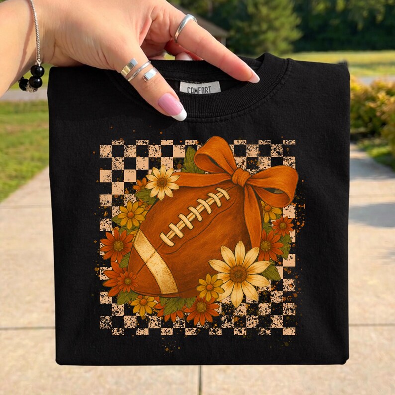 May include: Black sweatshirt featuring a football design with a bow, surrounded by flowers, set against a checkered background. The football is brown with white laces. The word "COMFORT" is printed on the collar.
