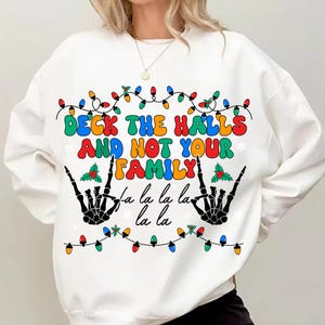 Op de afbeelding: Witte sweatshirt met de tekst "Deck the Halls and Not Your Family" in kleurrijke letters. Het ontwerp bevat zwarte skelet handen, kerstverlichting en hulst accenten. Een feestelijk grafisch ontwerp met een vakantiethema.