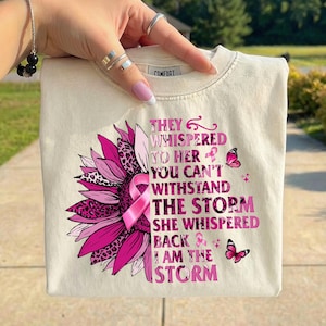May include: Cream-colored sweatshirt featuring a pink sunflower design with a leopard print center and a pink ribbon. The text reads "THEY WHISPERED TO HER YOU CAN'T WITHSTAND THE STORM SHE WHISPERED BACK I AM THE STORM".