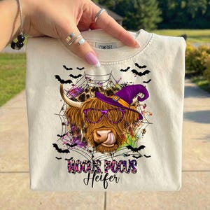 May include: A cream-colored t-shirt featuring a graphic of a Highland cow wearing a purple witch hat and purple leopard-print glasses. The cow is set against a black spiderweb and bat design with colorful paint splatters. Text reads Hocus Pocus Heifer.