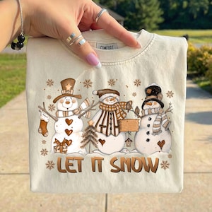 May include: Cream-colored sweatshirt featuring a winter scene with three snowmen. Each snowman has a unique hat, scarf, and accessories. The phrase "LET IT SNOW" is printed in brown script below the snowmen. The word "COMFORT" is printed on the collar.