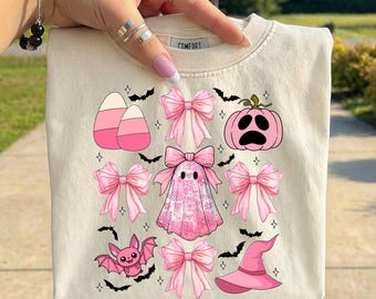 May include: A cream-colored t-shirt featuring a graphic print of pink Halloween-themed icons including a ghost, a pumpkin, a bat, a witch hat, candy corn, and several bows. The design is arranged in a grid pattern with small black bats and stars.