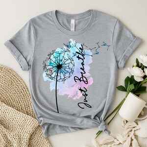 May include: A light gray t-shirt featuring a dandelion design with the words "Just Breathe" in black script. The dandelion is accented with blue and purple watercolor and butterflies.