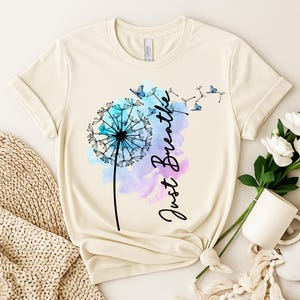 May include: Cream-colored t-shirt featuring a dandelion design with blue and purple watercolor accents. The words "Just Breathe" are written in black script. The dandelion's seeds transform into butterflies.