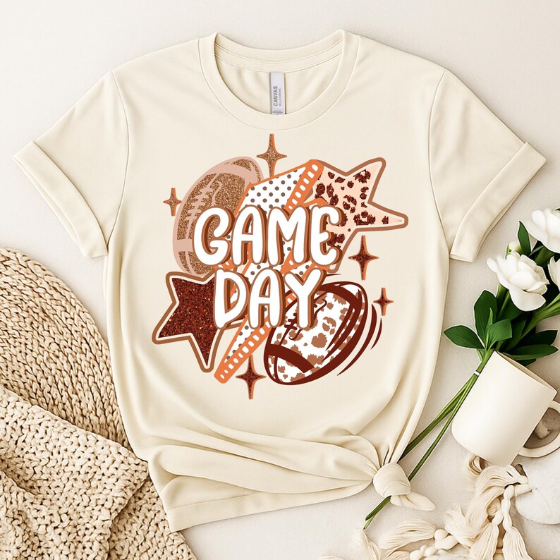 May include: Cream-colored t-shirt featuring a "GAME DAY" graphic with footballs and star shapes. The design incorporates glitter, leopard print, and brown and orange accents. The shirt has rolled-up sleeves and a tied knot at the bottom.