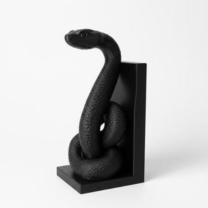 Op de afbeelding: Een zwarte slangenbeeldhouwwerk, opgerold en rechtop, tegen een zwarte rechthoekige steun. De slang heeft een getextureerd oppervlak en een gedetailleerde kop. De sculptuur staat tegen een witte achtergrond.