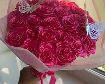 May include: A bouquet of vibrant fuchsia roses, wrapped in pink paper and tied with a matching ribbon. Two decorative white butterfly cutouts are attached to the bouquet, adding a delicate touch. The bouquet is held by a person.