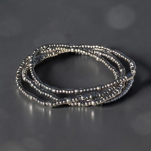 May include: A set of silver beaded bracelets. The bracelets are made of small, round beads strung together. The bracelets are coiled together on a gray surface, creating a layered effect. The beads reflect light, giving them a shiny appearance.