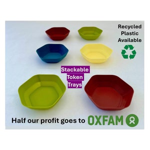 May include: Five hexagonal plastic trays in different colours: green, blue, yellow, red, and red. The trays are stackable and made from recycled plastic. The text "Stackable Token Trays" is written in purple letters. The text "Recycled Plastic Available" is written in black letters. The text "Half our profit goes to OXFAM" is written in black letters. A green recycling symbol is shown.