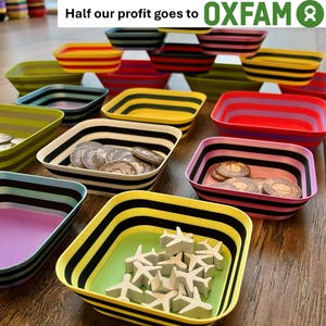 May include: A collection of square, multi-coloured plastic bowls with horizontal striped patterns. Some bowls contain small, round metallic coins or white aeroplane-shaped tokens. The text "Half our profit goes to OXFAM" is visible at the top.