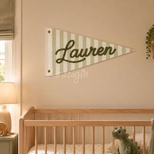 Op de afbeelding: Een driehoekige wandwimpel met een lichtgroen en wit verticaal gestreept patroon en de naam Lauren in donkergroen cursief schrift. De wimpel is voorzien van twee metalen ringen aan de linkerkant om aan de muur op te hangen.