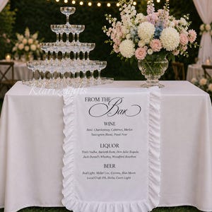 Può includere: Allestimento bar per ricevimento di nozze con tovaglia bianca, torre di bicchieri di champagne e composizione floreale. Un runner bianco con il testo "FROM THE Bar" elenca le opzioni di vino, liquore e birra.