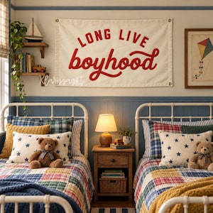 Può includere: Uno stendardo in tessuto color crema con il testo rosso 'LONG LIVE boyhood' appeso sopra due letti singoli in metallo bianco. Ogni letto presenta una trapunta a quadri, un cuscino bianco con stelle blu e un orsacchiotto di peluche marrone.