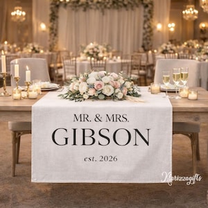 Könnte beinhalten: Eine Hochzeitstafel mit einem weißen Tischläufer. Der Läufer trägt den Text "MR. & MRS. GIBSON est. 2026" in Schwarz. Ein Blumengesteck aus weißen und rosa Rosen liegt obenauf. Sektgläser und Kerzen stehen ebenfalls auf dem Tisch.