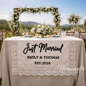 May include: A wedding reception table is set with a linen tablecloth and a decorative lace runner. The runner features the words "Just Married", the names "Emily & Thomas", and the year "EST.2026". A floral arrangement and glassware are on the table.