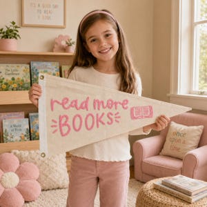 Puede incluir: Un banderín triangular de lona color crema con el texto bordado en rosa "read more BOOKS" y un pequeño icono de libro rosa. El banderín tiene una franja vertical blanca en el lado izquierdo con dos ojales metálicos de color latón.
