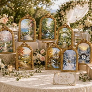 Könnte beinhalten: Eine Sammlung von acht gewölbten Tischnummernschildern in verzierten Goldrahmen, jedes mit einem einzigartigen Landschaftsgemälde und dem Text Table 1 bis 8, sowie einem rechteckigen Schild Cards & Gifts, alles auf einer weißen Tischdecke.