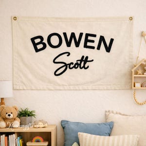 May include: Cream-coloured banner with the name "BOWEN Scott" in black, bold, and cursive lettering. The banner has brass eyelets in the corners for hanging. The banner is displayed on a white wall above a shelf with a teddy bear and books.