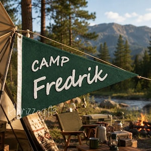 May include: A triangular dark green felt pennant featuring the white embroidered text "CAMP Fredrik" in a mix of block and script lettering. The pennant has a white vertical strip along the left edge with two metal grommets for hanging.