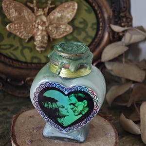 Può includere: Una bottiglia a forma di cuore blu chiaro con un sigillo di cera verde. La bottiglia presenta un adesivo a forma di cuore con un bordo nero e un'immagine in bianco e nero di Frankenstein e la sua sposa. Una falena dorata sullo sfondo.