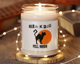 Może przedstawiać: Zapalone świeca w szklanym słoiku z etykietą "BLACK CAT FULL MOON". Etykieta przedstawia sylwetkę czarnego kota na tle pomarańczowego koła. Świeca stoi na drewnianej podstawce, z łańcuchem świetlnym na pierwszym planie.