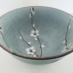 May include: A round, pale blue ceramic bowl with a brown rim. The interior features a delicate floral design of white blossoms and black stems. The bowl has a textured appearance, suggesting a handcrafted quality. Suitable for serving or display.