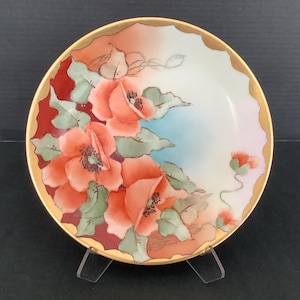 May include: A decorative plate with a gold rim and a floral design. The plate features orange poppies with green leaves against a backdrop of red, blue, and peach hues. The plate is displayed on a clear stand. The plate's diameter is approximately 20 cm.