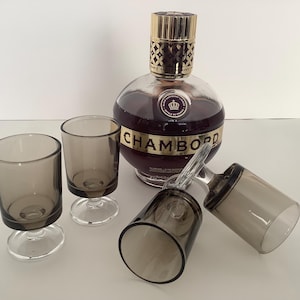 May include: A bottle of Chambord liqueur with a gold cap and label, accompanied by four gray-tinted shot glasses. The bottle is round and filled with a dark red liquid. The glasses are clear with a stem.