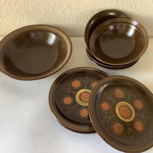 Pode incluir: Um conjunto de louças de cerâmica marrom, incluindo tigelas e pratos. Os pratos apresentam um padrão decorativo com detalhes em laranja e dourado. O conjunto inclui uma tigela grande e várias tigelas e pratos menores.