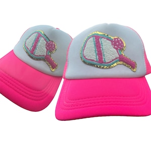 May include: Two pink and white trucker hats with a white and pink paddleball design on the front. The paddleball design has a gold outline.