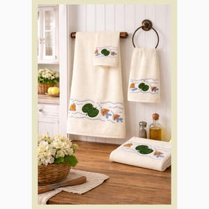 May include: A set of cream-colored bath towels with embroidered aquatic designs. The towels feature a pattern of green lily pads, orange fish, and blue waves. The set includes a bath towel, hand towel, and washcloth.