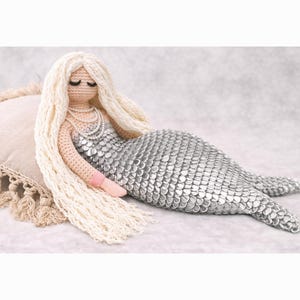 May include: A handmade crochet mermaid doll with long, flowing blonde hair and a silver scale tail. The mermaid is wearing a pearl necklace and has pink accents on her arms. The doll is lying on a beige pillow.
