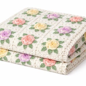 May include: Folded cream-colored crochet blanket with a floral pattern. The blanket features squares with embroidered roses in pink, yellow, and purple, surrounded by green leaves. The blanket is folded in half.