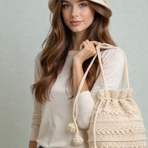 May include: A cream-colored crochet bucket hat and drawstring bag. The hat has a rounded crown and a small brim. The bag features a textured stitch pattern and pom-pom accents. The bag has a drawstring closure and long straps.