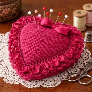 Może przedstawiać: Poduszka na szpilki w kształcie serca, w kolorze magenta, z szydełkową fakturą i falistą satynową obwódką. Kilka szpilek z kolorowymi główkami jest włożonych w poduszkę. W tle akcesoria do szycia, w tym szpulki nici i nożyczki.