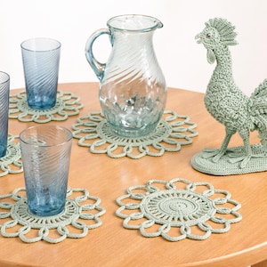 May include: A table setting featuring blue-tinted drinking glasses, a clear glass pitcher, and a crocheted rooster figurine. The glasses rest on matching crocheted doilies. The items are arranged on a round wooden table.