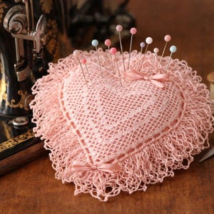 May include: A pink, heart-shaped crochet pin cushion with a frilled edge, holding pins with coloured heads. A vintage sewing machine, thread spool, and scissors are also in the image.