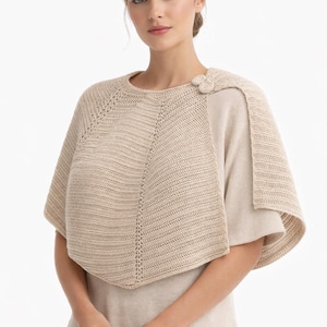 Puede incluir: Una capa de punto beige con cuello redondo y cierre de botón decorativo. La capa presenta un diseño texturizado y se usa sobre una blusa de color claro, creando un look en capas. La prenda está hecha de un material suave y de aspecto natural.