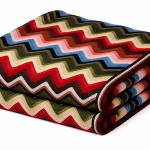 Puede incluir: Manta plegada con un colorido patrón de zigzag. La manta presenta un diseño repetitivo de zigzags rojos, blancos, verdes, marrones, azules y rosas. Los bordes están acabados con un borde marrón oscuro.