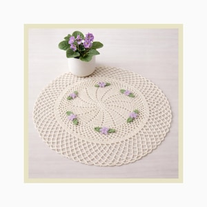 Może przedstawiać: Dwie kremowe, szydełkowe serwetki, jedna na drugiej, ze spiralnym wzorem i kwiatowymi akcentami. Górna serwetka ma małe fioletowe kwiaty i zielone liście. W pobliżu stoi biała doniczka z rośliną fiołka.