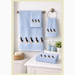 May include: A light blue, embroidered penguin-themed towel set. The set includes a bath towel, hand towel, and washcloth, each featuring a row of penguins. The towels are displayed in a bathroom setting.