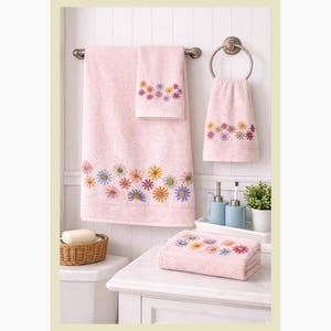 May include: A set of pink bath towels with embroidered colorful flowers. The set includes a large bath towel, a hand towel, and a washcloth. The towels are displayed in a bathroom setting with a soap dish and decorative items.