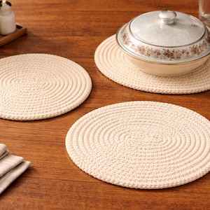 May include: Cream-colored, round placemats with a woven texture are displayed on a wooden table. A floral-decorated serving bowl with a lid sits on one of the placemats. A wooden spoon and folded napkins are also present.