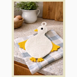 May include: A white crochet duck-shaped hot pad with yellow wings and feet, resting on a blue and white checkered towel. The duck has a yellow beak and a black eye. The background includes a white pitcher, a basket of eggs, and a wooden cutting board.