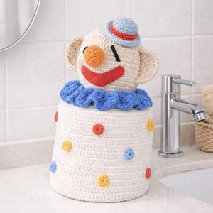May include: A handmade, crocheted clown-themed toilet paper cover. The cover is white with colorful polka dots and a clown face on top, complete with a small hat. The clown has a red smile, orange nose, and blue ruffled collar.
