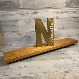 May include: A gold-colored letter N standing upright on a long, rectangular wooden shelf. The letter N features the name Natalie written in white cursive script vertically along its right side. The shelf has a natural wood grain finish.