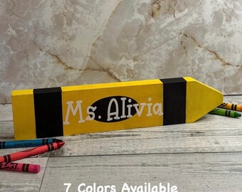 May include: A yellow and black wooden pencil-shaped sign featuring the white text Ms. Alivia printed on a black oval background. The sign is positioned horizontally with several colorful crayons scattered nearby on a light-colored wooden surface.