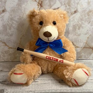 May include: A light brown plush teddy bear with a bright blue satin bow around its neck. The bear holds a miniature wooden baseball bat featuring the red text COLTON #5. The bear's feet have white patches with red stitching resembling a baseball.