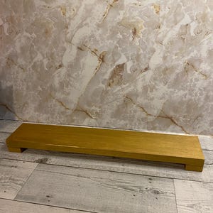 May include: A rectangular wooden riser with a smooth, metallic gold finish. The platform is elevated by two short, block-style feet on either end, creating a low-profile stand suitable for displaying various items on a flat surface.